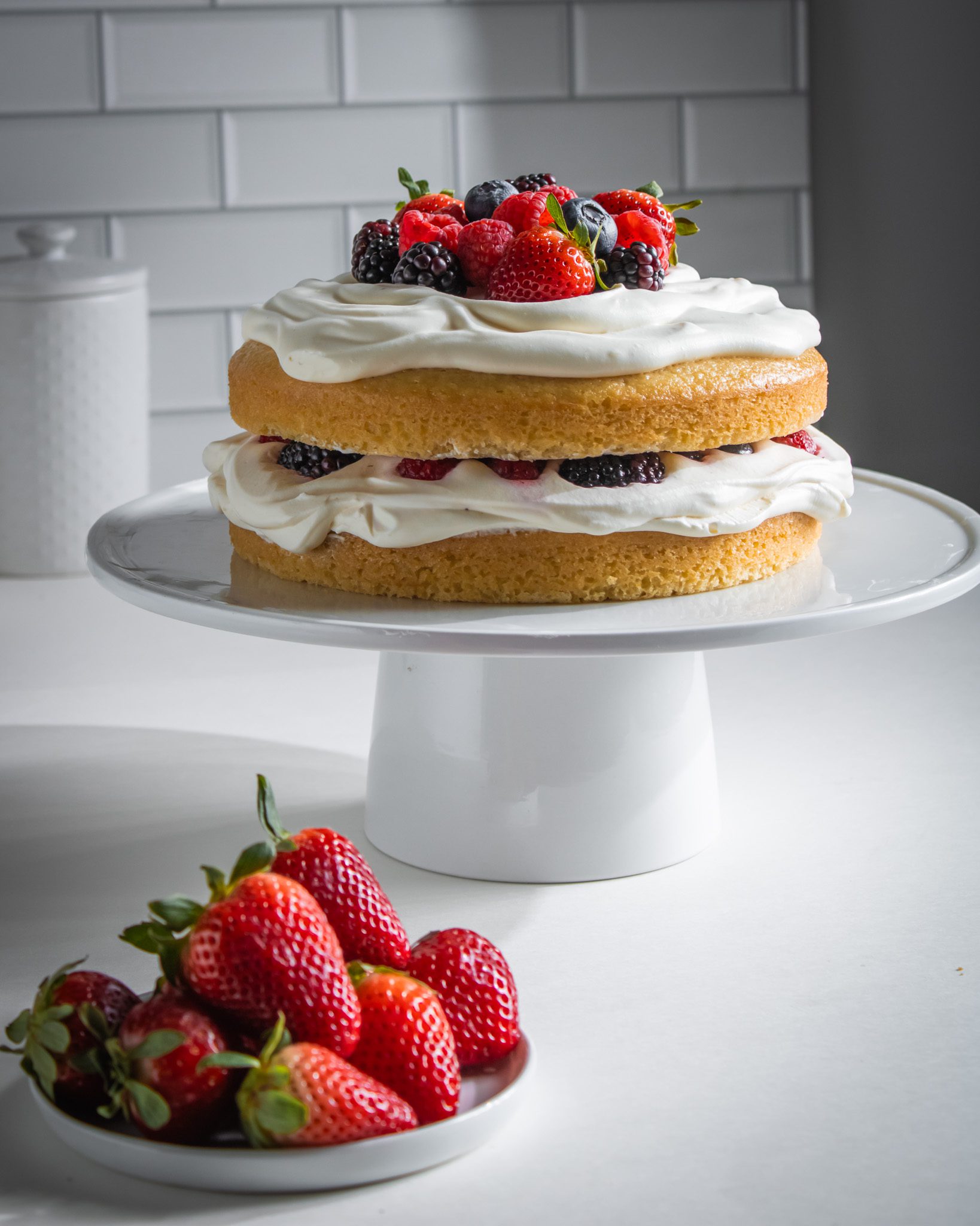 Eggless berry cream cake topped with fresh strawberries, blueberries, raspberries, and whipped cream.